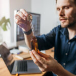 Man holding a dropper and amber bottle of broad spectrum CBD oil at his desk, examining the product closely.