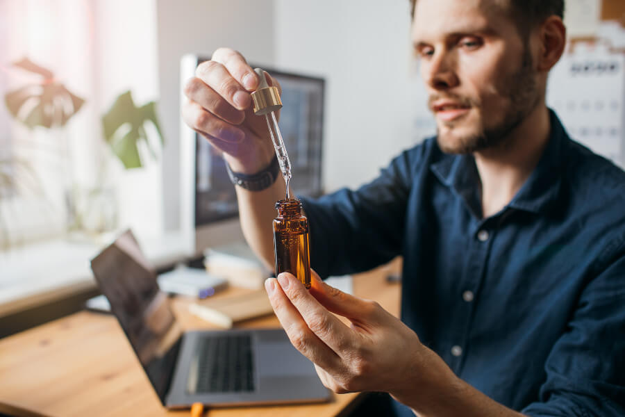 Man holding a dropper and amber bottle of broad spectrum CBD oil at his desk, examining the product closely.