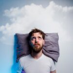 Tired man with dark under-eye circles lying awake in bed with a cloud above him, representing sleep problems and the potential benefits of CBD oil.