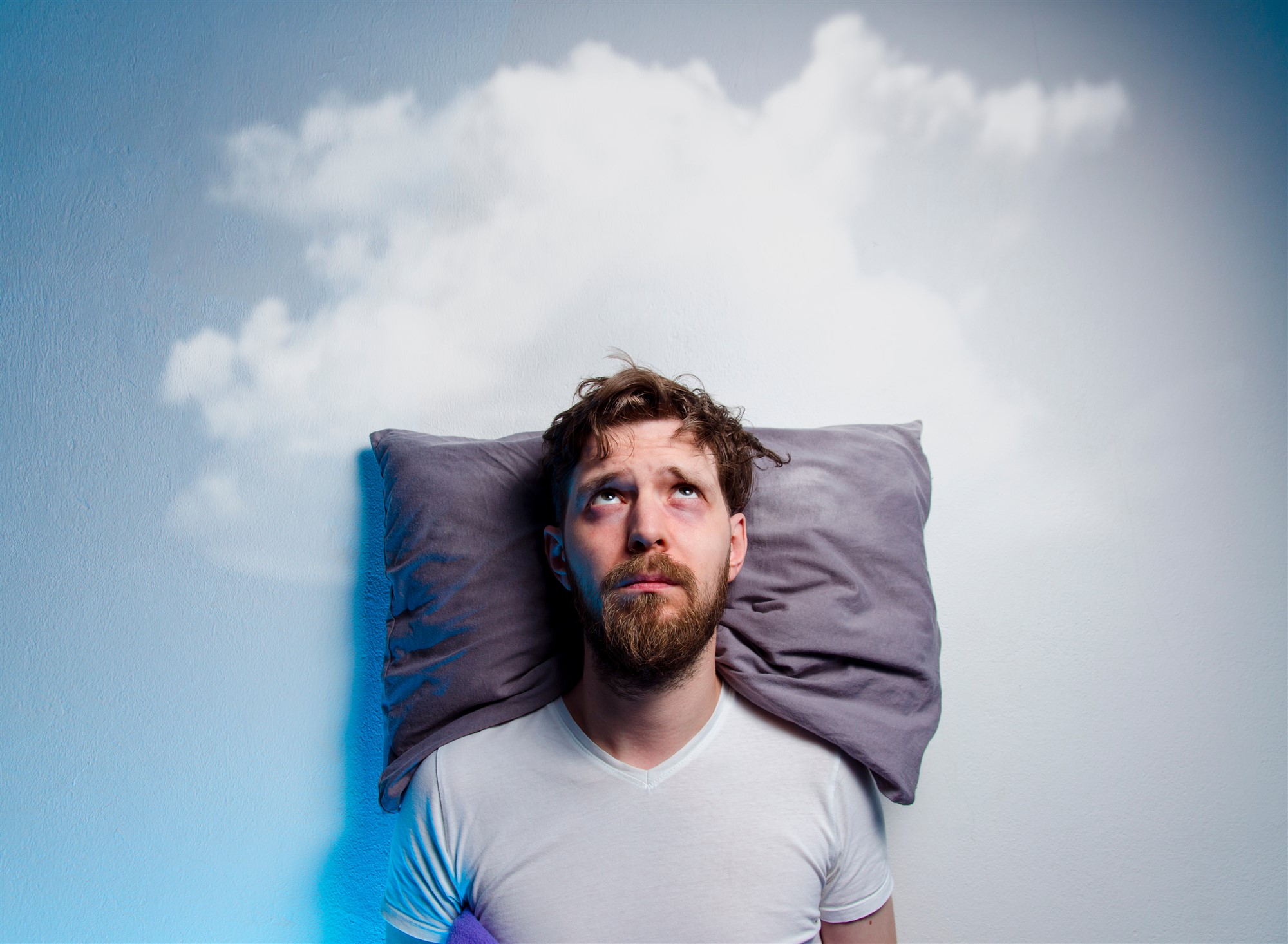 Tired man with dark under-eye circles lying awake in bed with a cloud above him, representing sleep problems and the potential benefits of CBD oil.